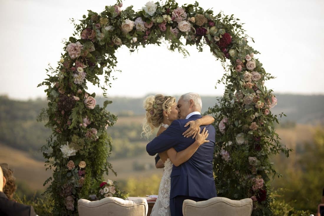 A couple embrace under a garden-rose arch at the end of their civil ceremony, rolling hills behind, Il Mio Matrimonio