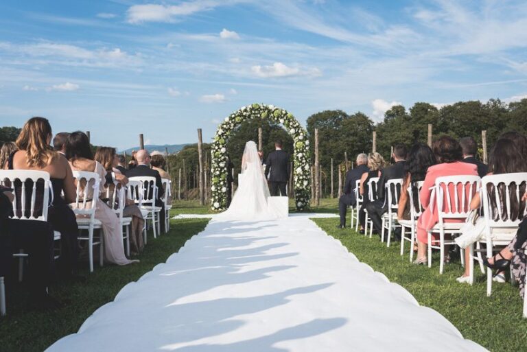 A white aisle runner across a lawn to a white-flower arch, guests on white chairs either side (Amalia and Sergio's wedding), Il Mio Matrimonio