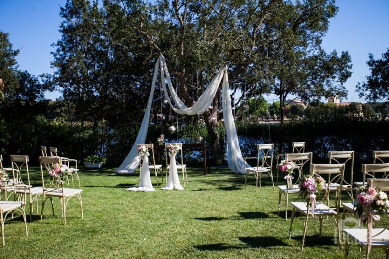 A garden ceremony set-up: a draped white fabric arch under the trees, chairs dressed with pink flowers (Daniele and Valentina's wedding), Il Mio Matrimonio