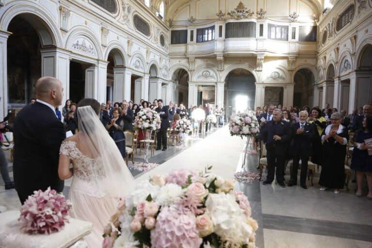 The bride at the head of the aisle in a baroque church, the pews lined with pink and white flowers (Eugenio and Simona's wedding), Il Mio Matrimonio