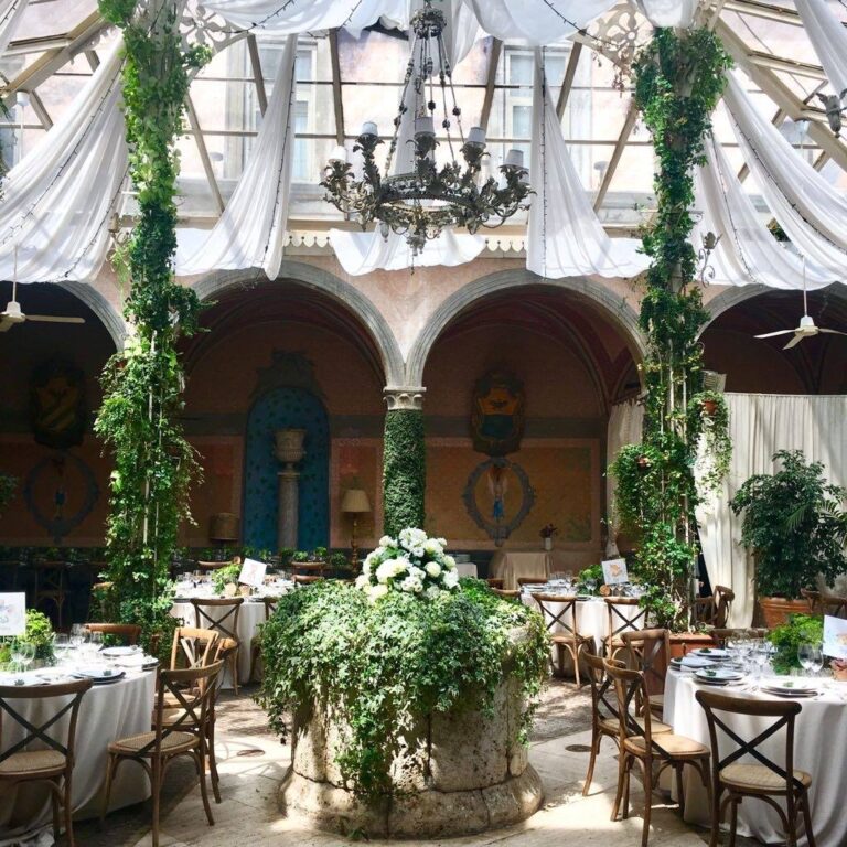 A glass-roofed arcaded courtyard set for dinner: round tables, wooden cross-back chairs and an ivy-covered well at the centre, Il Mio Matrimonio