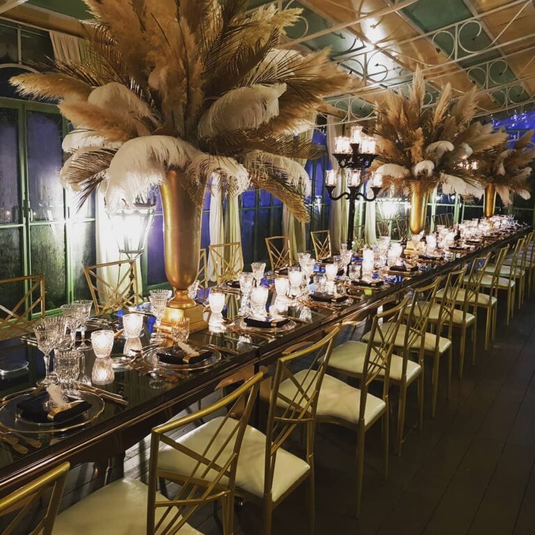 A long table under a glasshouse roof with tall pampas and feather centrepieces in gold vases and gold bamboo chairs, Il Mio Matrimonio