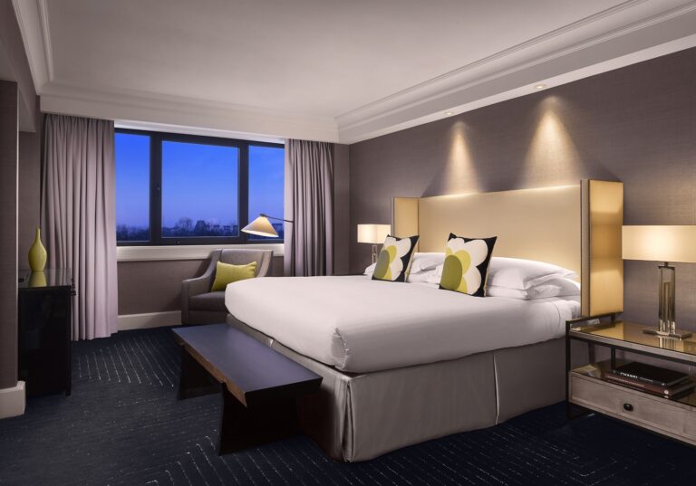 A Palace Suite bedroom at dusk, with a king bed, patterned carpet and windows over the park, InterContinental London Park Lane