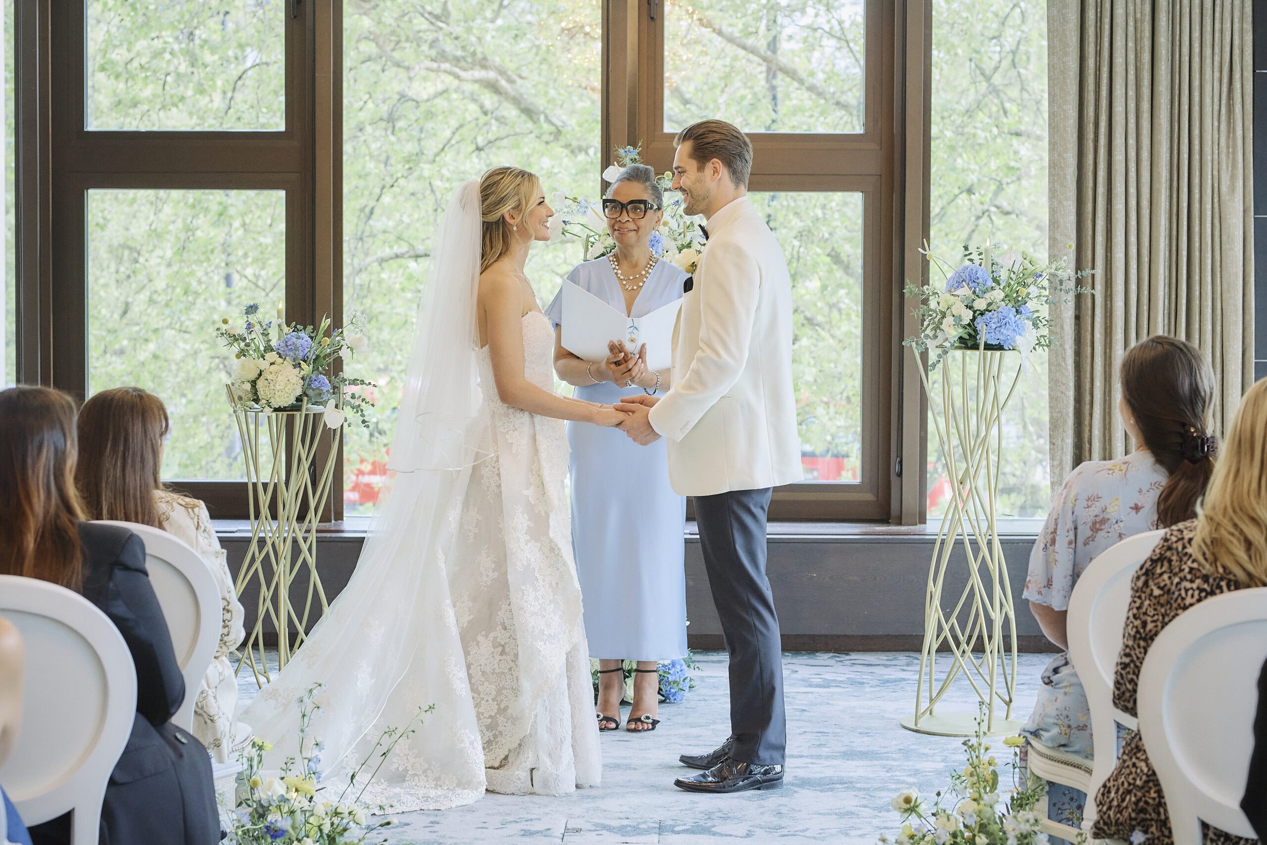 A couple holding hands before the celebrant at a small ceremony by the windows, with pastel flowers on gold stands, InterContinental London Park Lane