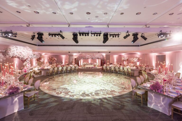 The Ballroom dressed for a large wedding in pink light, with a blossom arch over the top table, tables round a printed dance floor and full stage lighting, InterContinental London Park Lane
