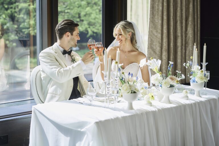 A couple toasting at a small top table dressed with white flowers and candles beside a window, InterContinental London Park Lane