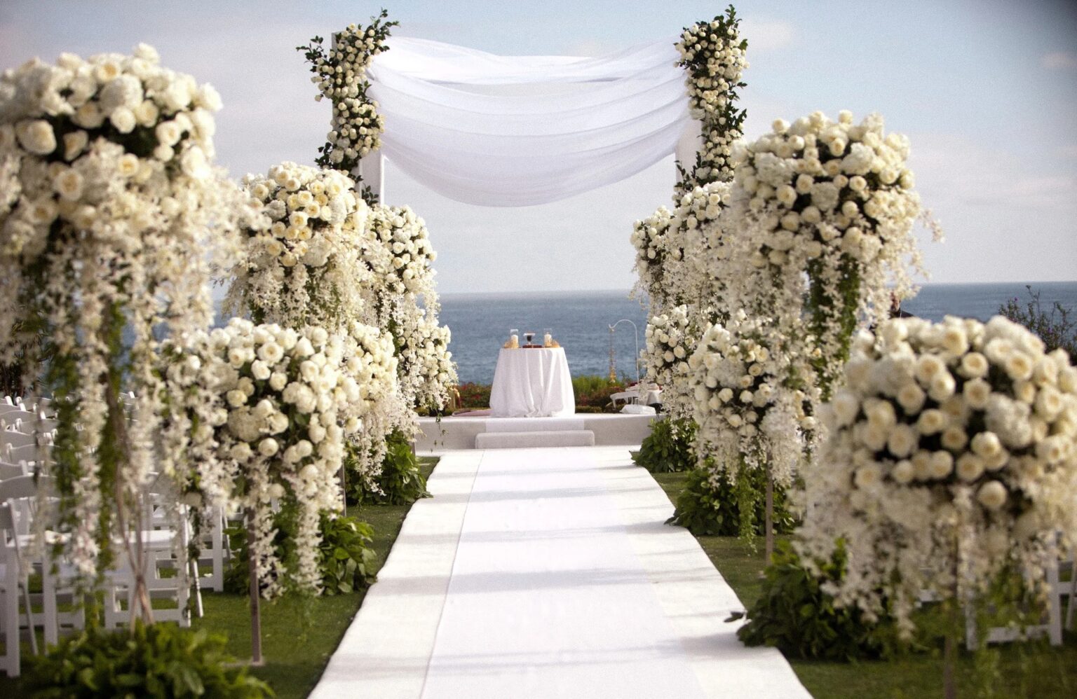 A white aisle between tall white flower columns to a draped canopy, with the sea beyond