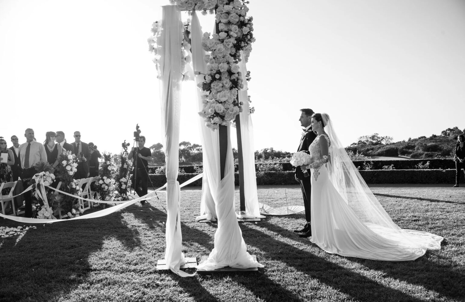 A couple at a draped ceremony canopy dressed with white flowers, in black and white, International Event Company