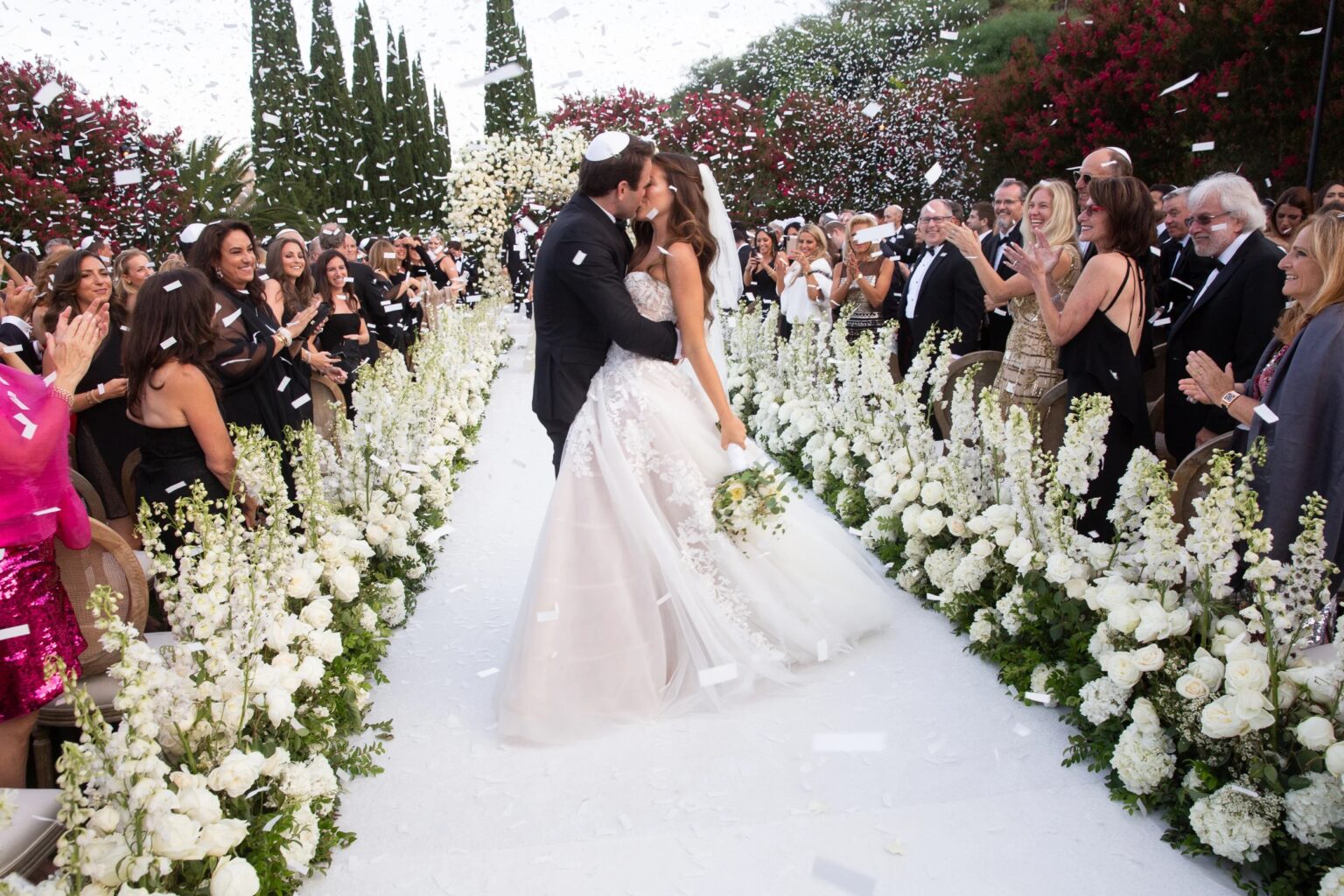 A couple kiss at the end of a white aisle lined with delphiniums as guests throw confetti