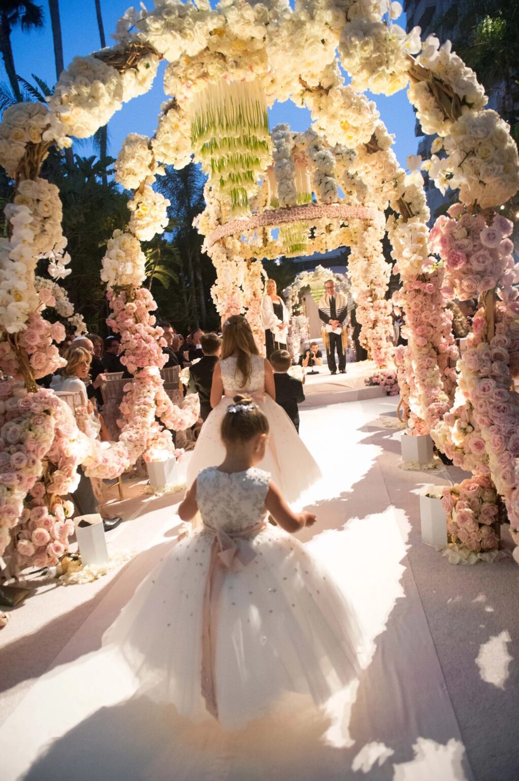 A flower girl walking beneath arches of white and blush roses with a hanging chandelier, International Event Company