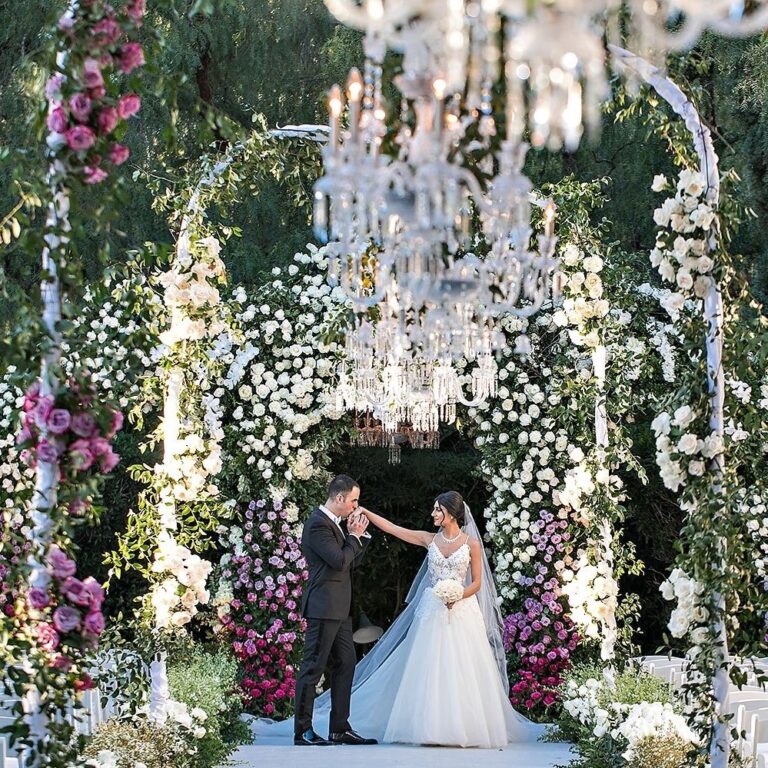 A groom kisses his bride's hand beneath a chandelier and an arch of roses, International Event Company