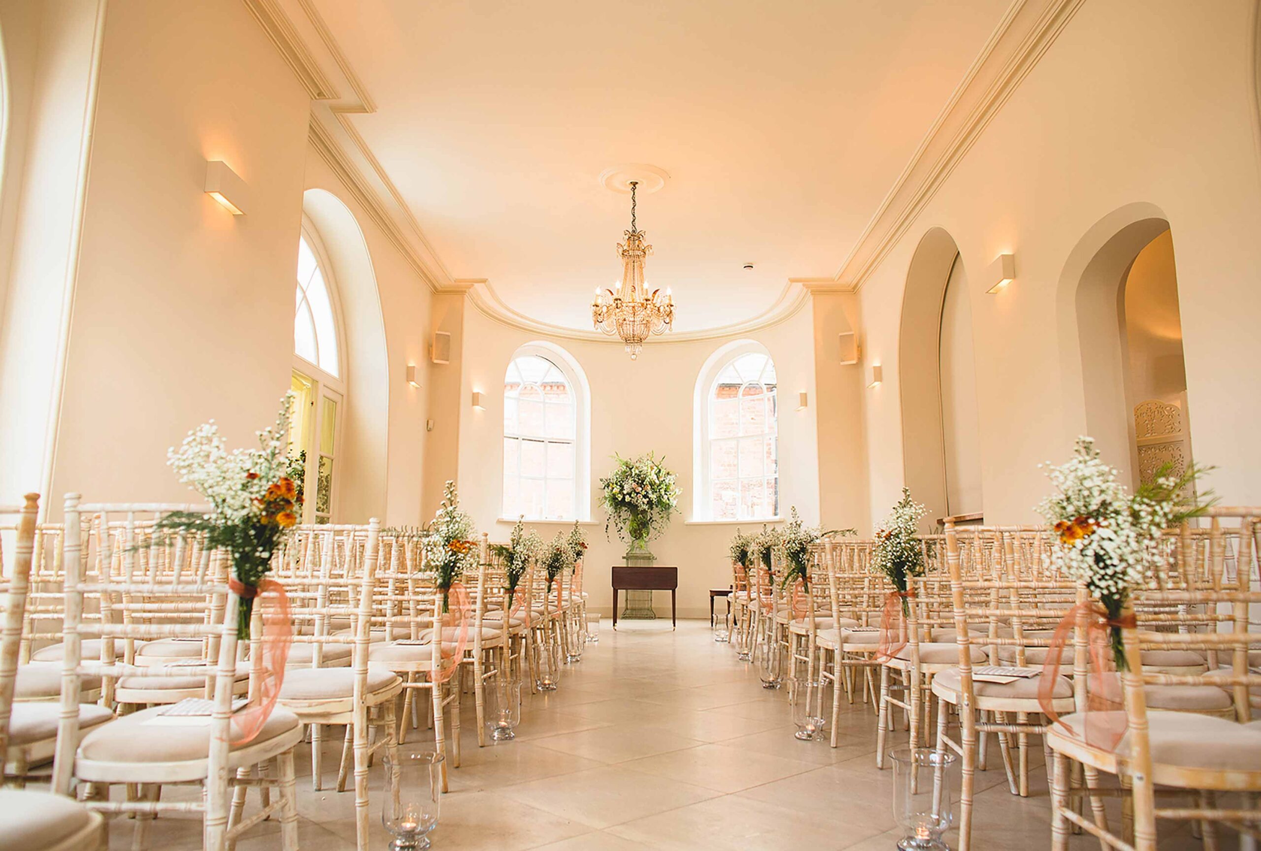 The Garden Room set for a ceremony, chairs in rows beneath a chandelier and tall arched windows, floral pedestals along the aisle, Iscoyd Park