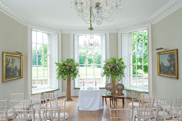 A light-filled drawing room with a bay window over the garden, set with chairs and floral pedestals for a ceremony, Iscoyd Park
