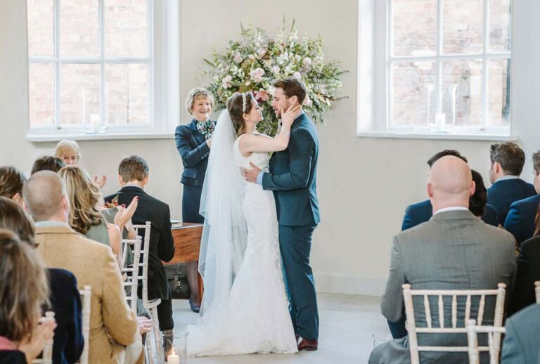 A couple kiss at their ceremony in the Garden Room beneath a floral arch and tall windows, Iscoyd Park