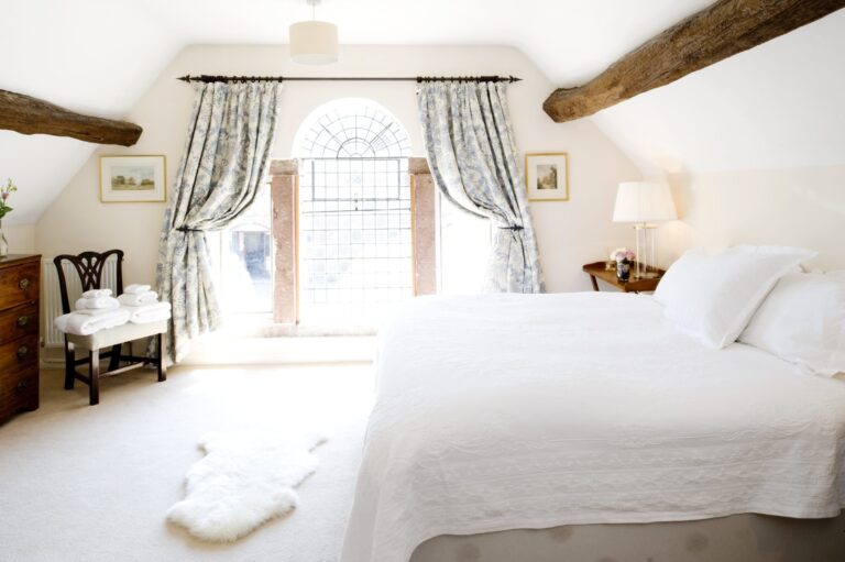 A bright, beamed bedroom with a white bed and an arched window framed by curtains, Iscoyd Park