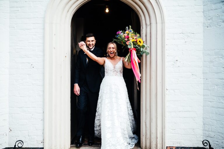 A bride and groom celebrate in the doorway of the house, a bouquet of bright wildflowers in hand, Iscoyd Park
