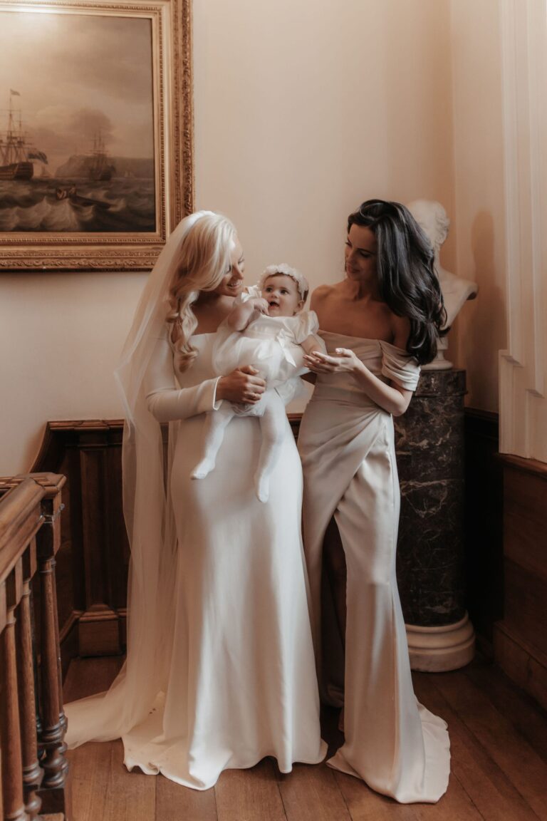 Two brides embrace in a room hung with gilt-framed oil paintings, Iscoyd Park