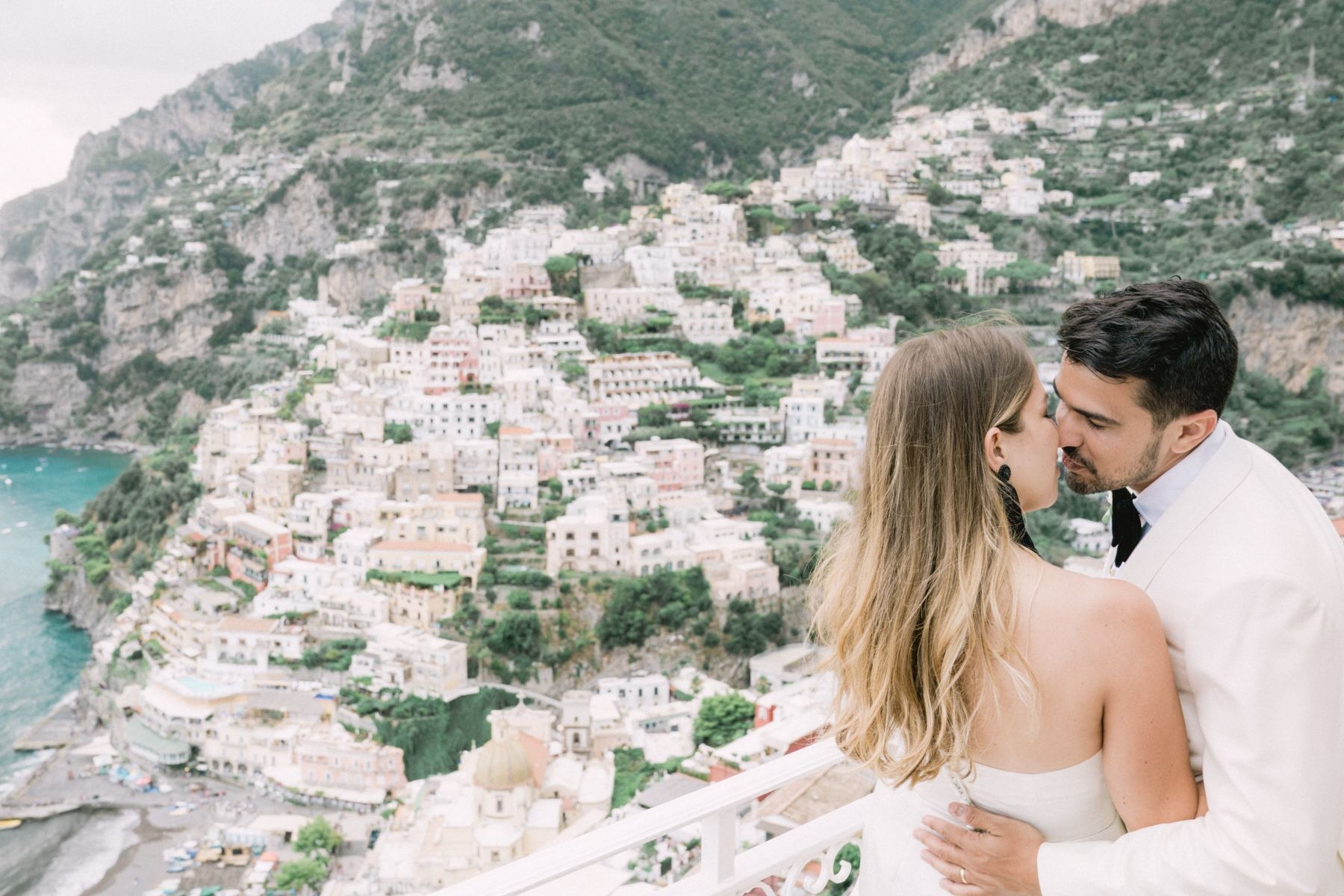 A couple kissing on a balcony above Positano, the pastel town stacked below, Italian Event Planners