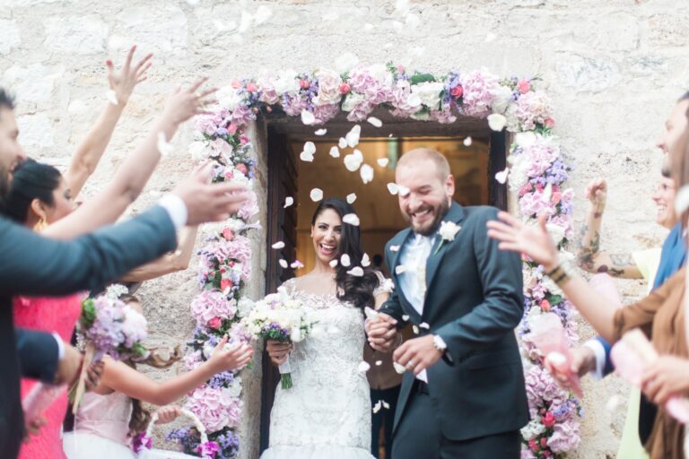 Guests throwing white petals over a couple in a flower-garlanded stone doorway, Italian Event Planners
