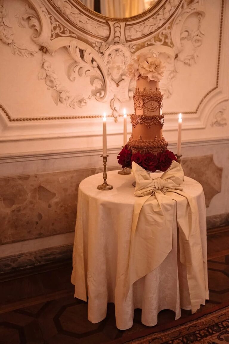 A tall ivory wedding cake with piped gold lattice on a round table, set against gilt stucco, Italian Eye