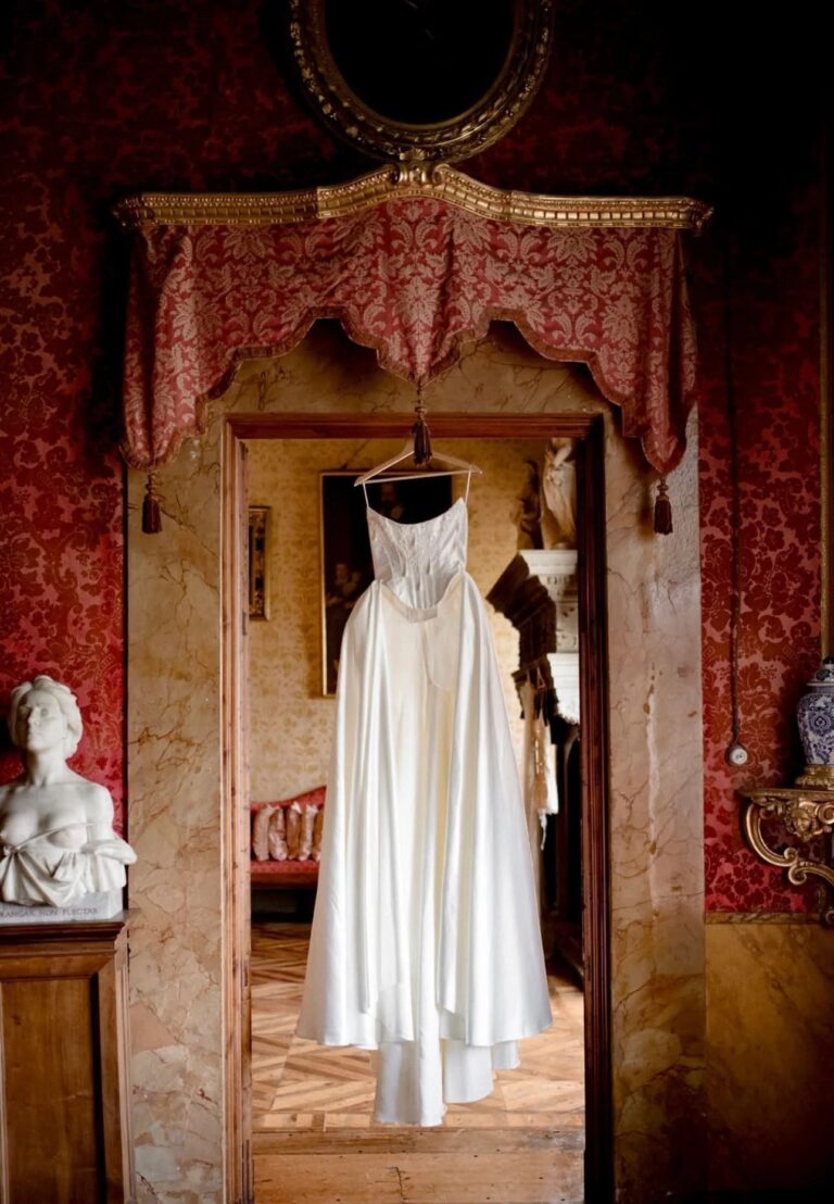 An ivory wedding gown hanging in a doorway between two damask-hung palazzo rooms, Italian Eye