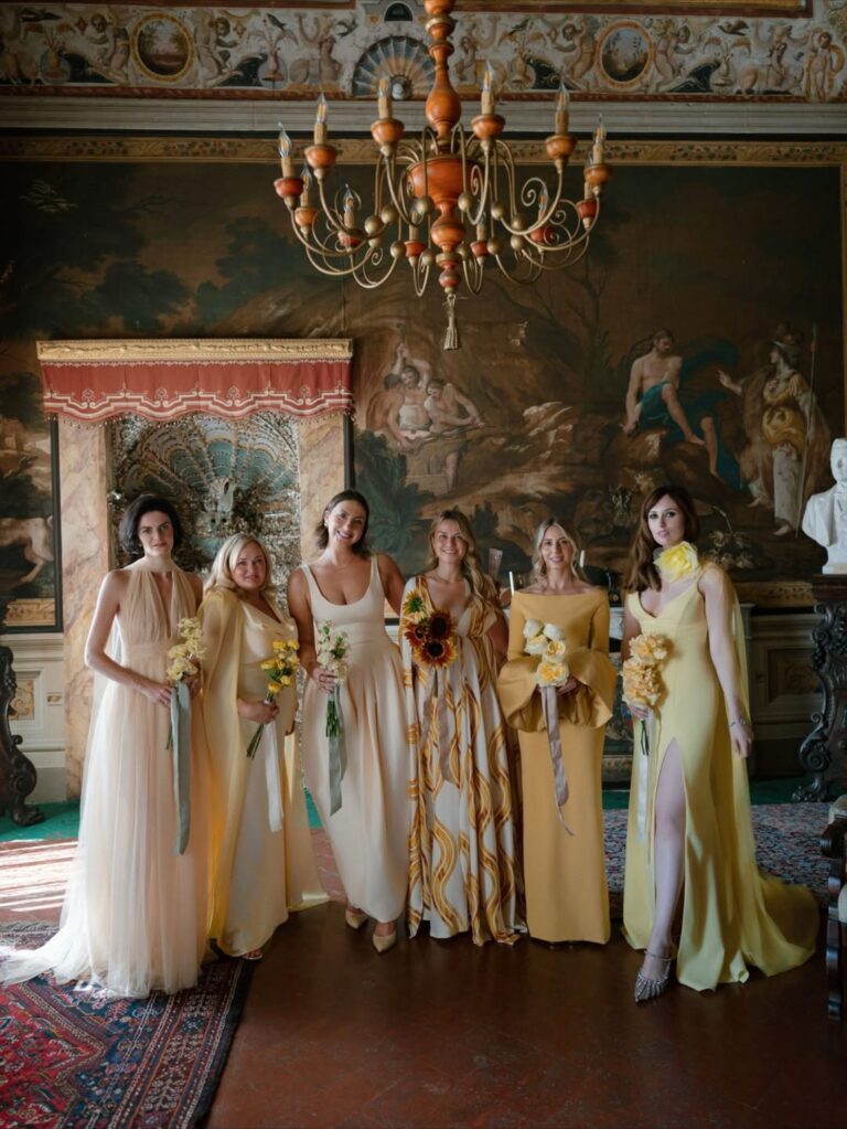 Bridesmaids in shades of cream and gold holding sunflowers beneath a chandelier in a frescoed palazzo room, Italian Eye