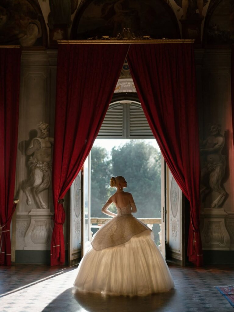 A bride in a full ballgown at an open window framed by red velvet curtains, Italian Eye