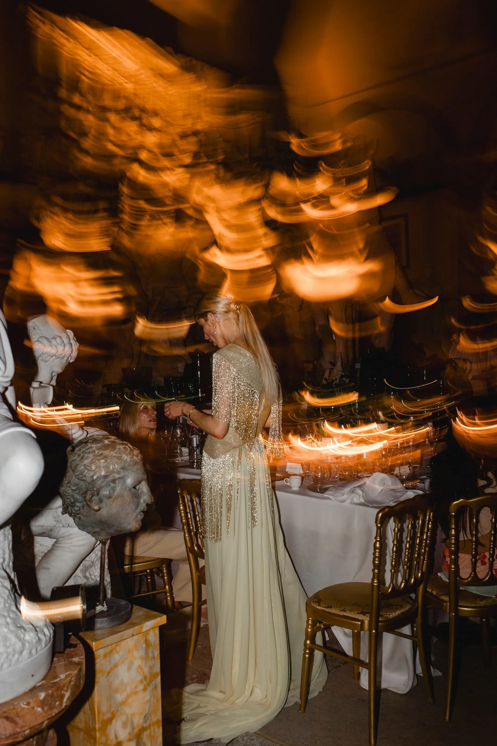 A reception in a Florentine sculpture gallery, a guest in a beaded gown caught in the candle light among marble busts, Italian Eye