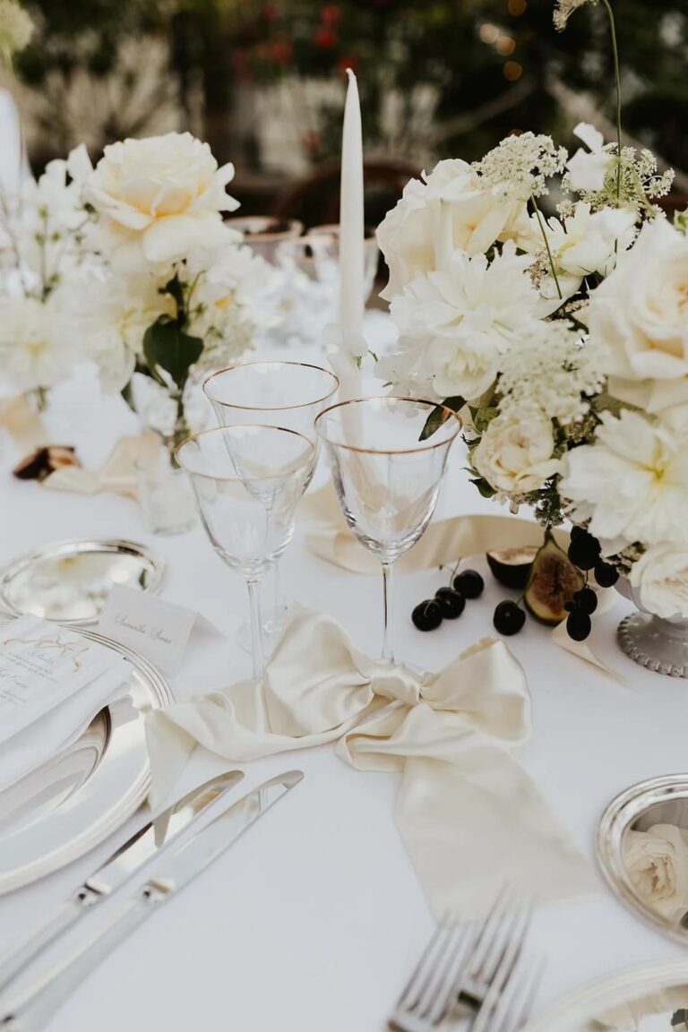 A white place setting with silver rimmed glasses, silk bow napkin and cream roses, Italian Eye