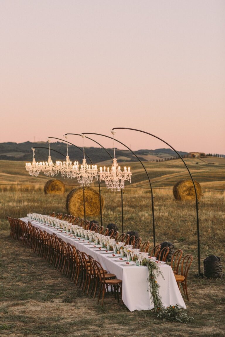 A long candlelit dinner table in a Tuscan field at dusk, five chandeliers on curved iron stands over hay bales, Italian Eye