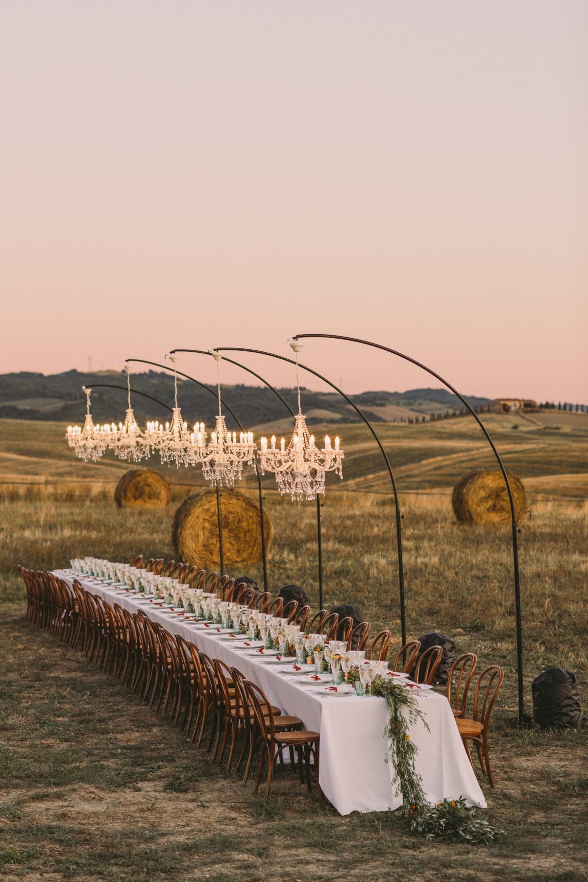A long candlelit dinner table in a Tuscan field at dusk, five chandeliers on curved iron stands over hay bales, Italian Eye