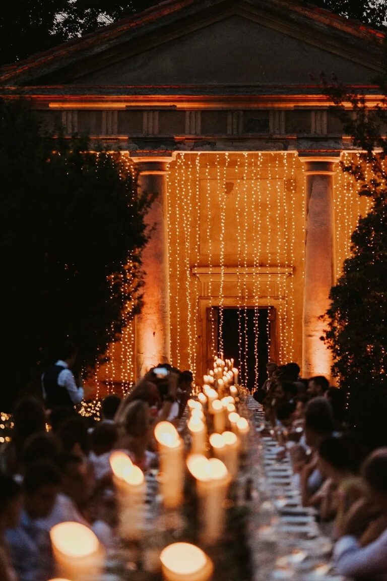 Guests at a long candlelit table facing a neoclassical temple strung with fairy lights, Italian Eye