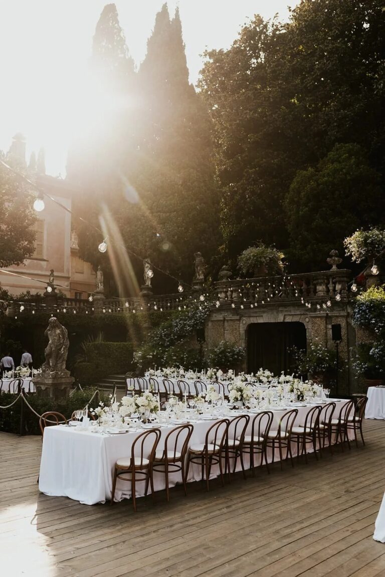 A sunlit terrace table beneath a balustrade of classical statues, with string lights overhead, Italian Eye