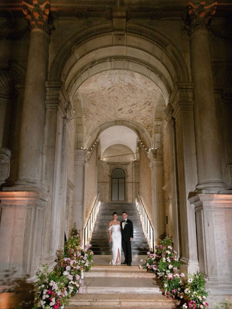 A couple at the foot of a stone staircase in a Venetian palazzo with floral garlands, Italian Eye