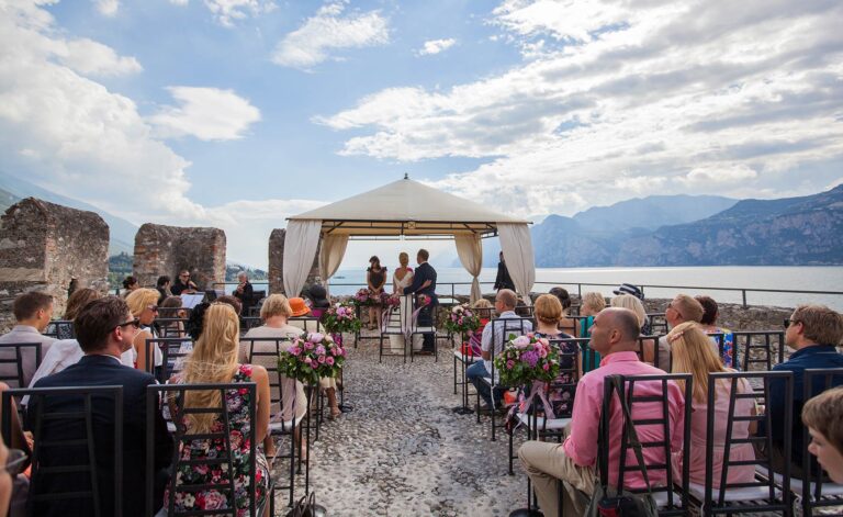 Guests seated on a castle terrace for a civil ceremony under a white canopy, the lake and mountains beyond, Italian Wedding Company