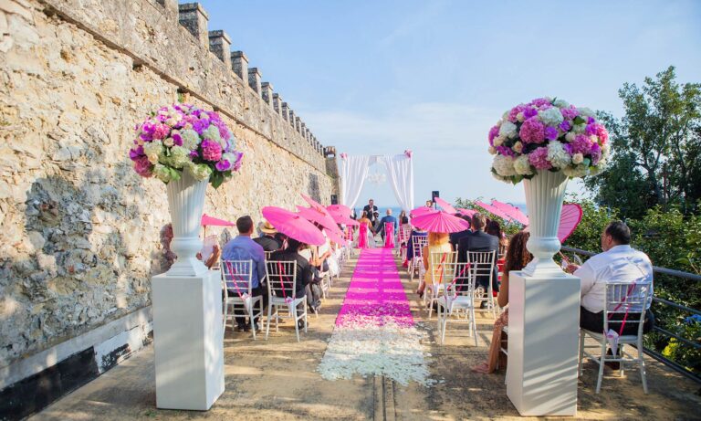 A symbolic ceremony on a castle terrace, pink parasols, a petal aisle and urns of peonies against the old walls, Italian Wedding Company