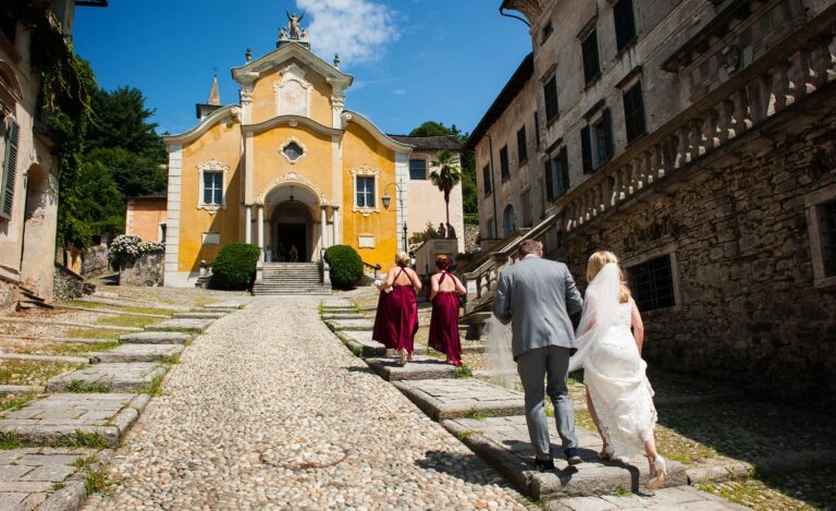 The bride on the cobbled lane up to the church of Santa Maria Assunta in Orta San Giulio, Lake Orta, bridesmaids in red ahead, Italian Wedding Company