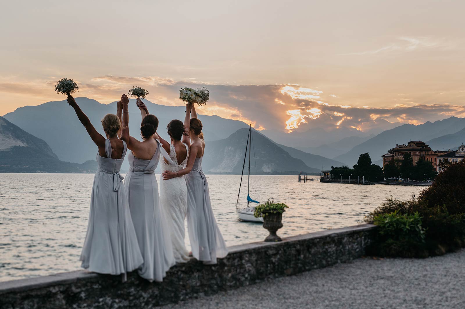 Bridesmaids raising their bouquets on the lakeside wall at sunset on Lake Maggiore, a villa on the shore beyond, Italian Wedding Company