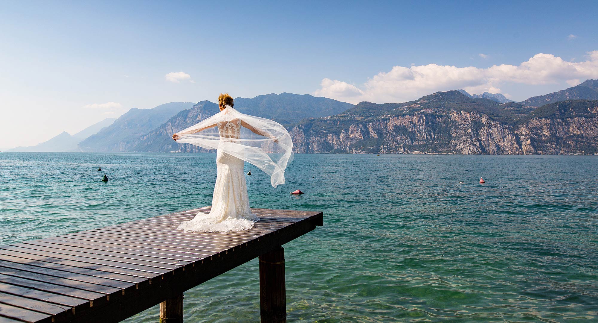 A bride alone at the end of a wooden jetty on Lake Garda, her veil lifting in the wind, the mountains of the far shore behind, Italian Wedding Company