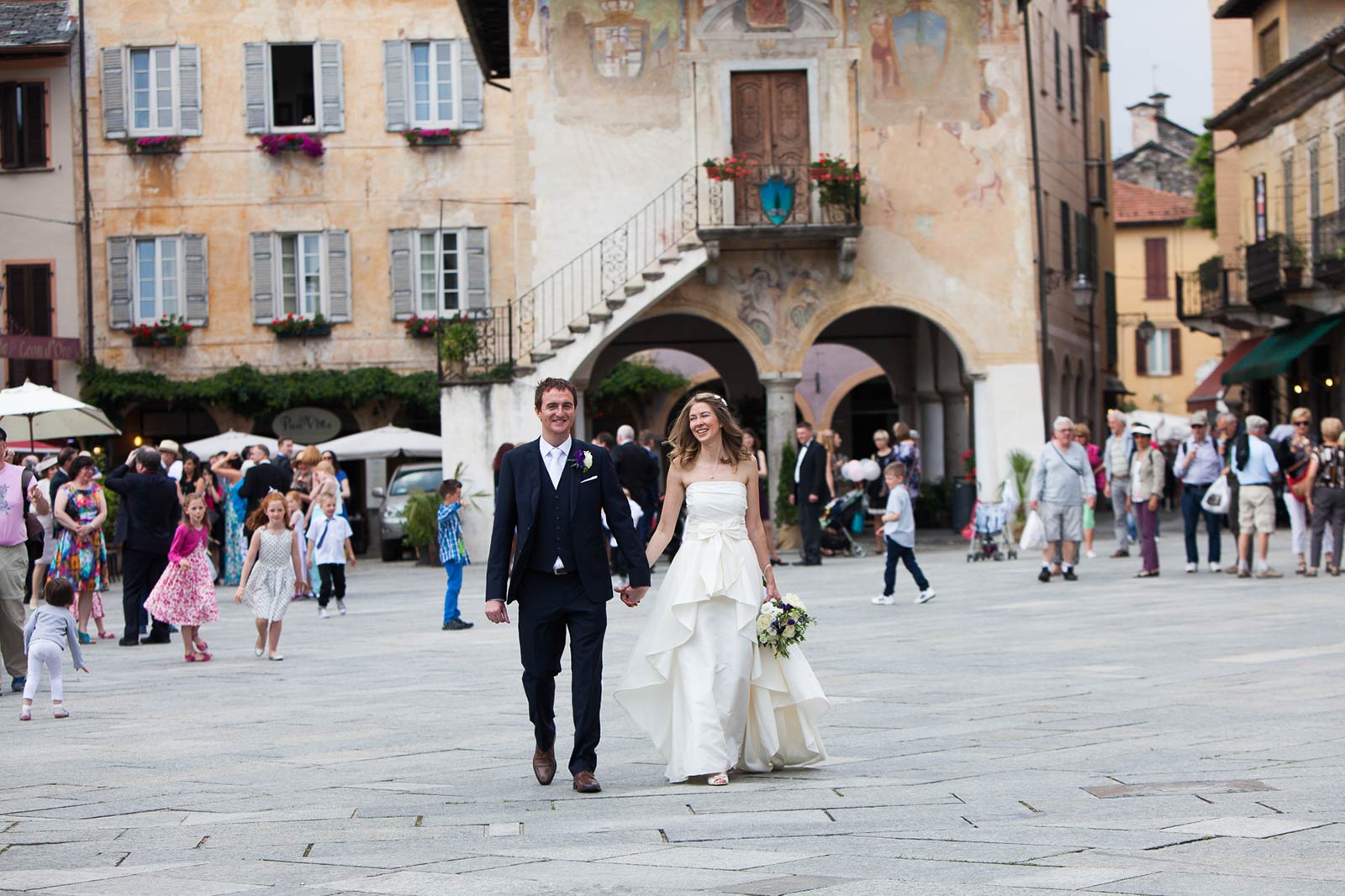 The couple walking out through Piazza Motta in Orta San Giulio, Lake Orta, guests and the painted palazzo behind them, Italian Wedding Company