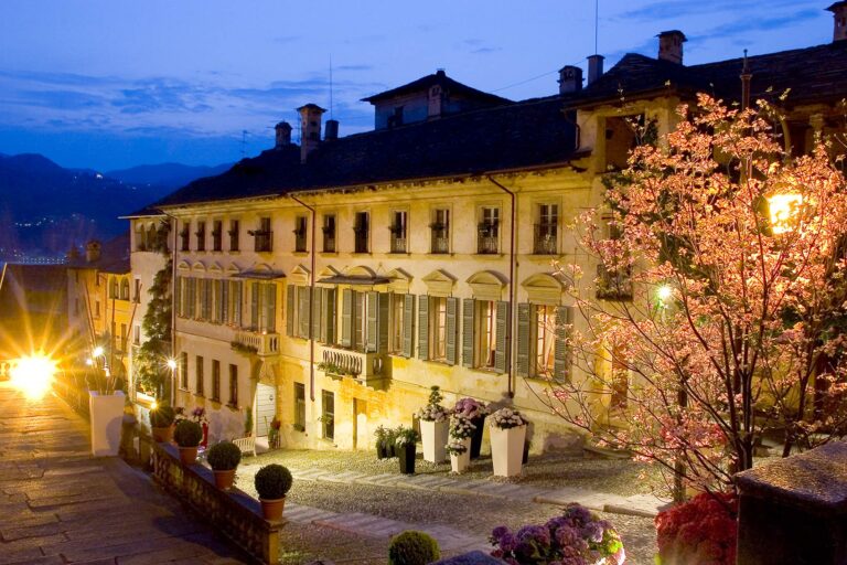 The facade and courtyard of Palazzo Penotti Ubertini in Orta San Giulio at dusk, a flowering tree lit against the stone, Italian Wedding Company