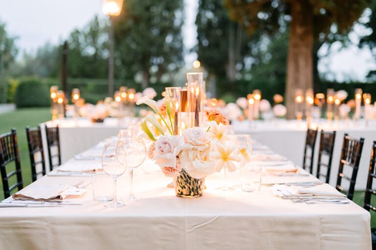 Long banquet table with peach flowers and candles under trees, Italian Wedding