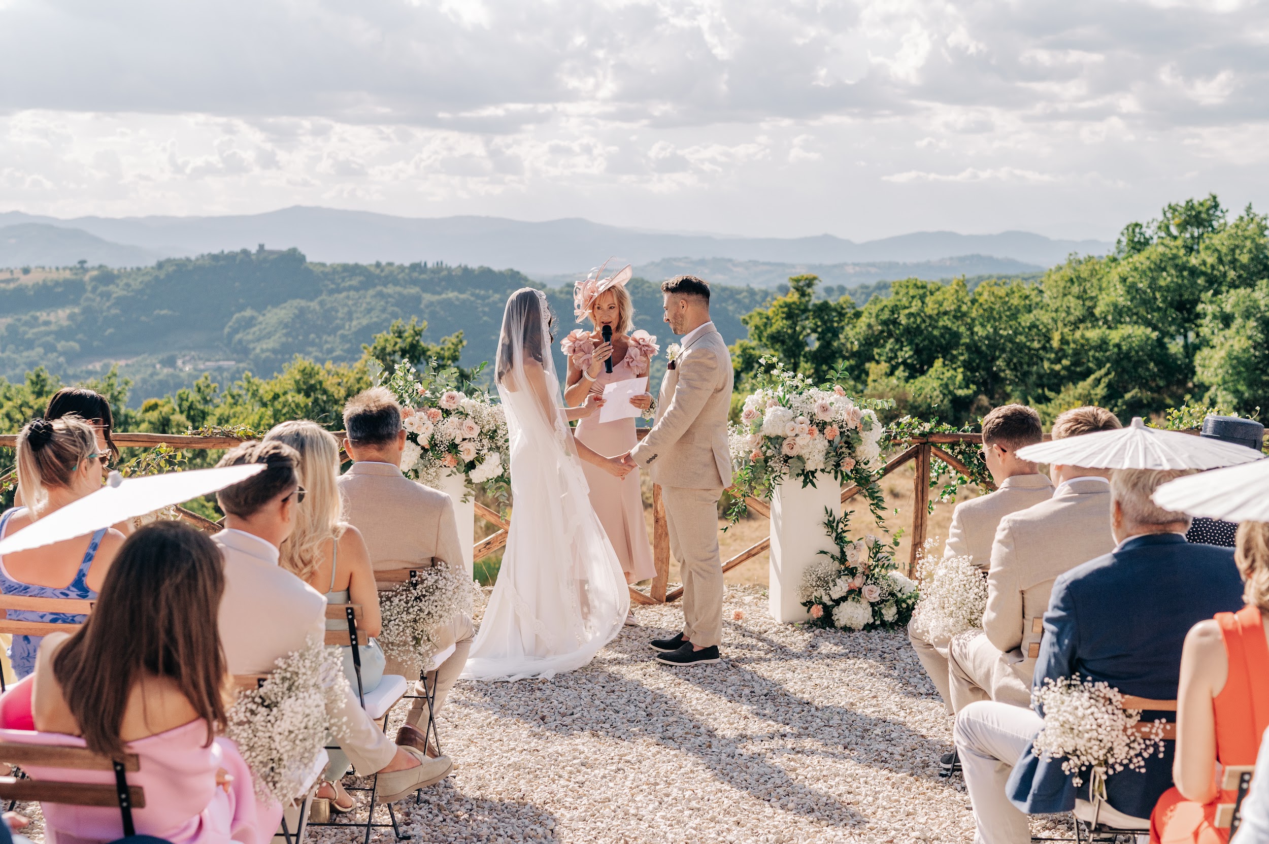 A ceremony on the terrace at Nikis Resort in Umbria, guests with white parasols and the hills behind, Joy Wedding