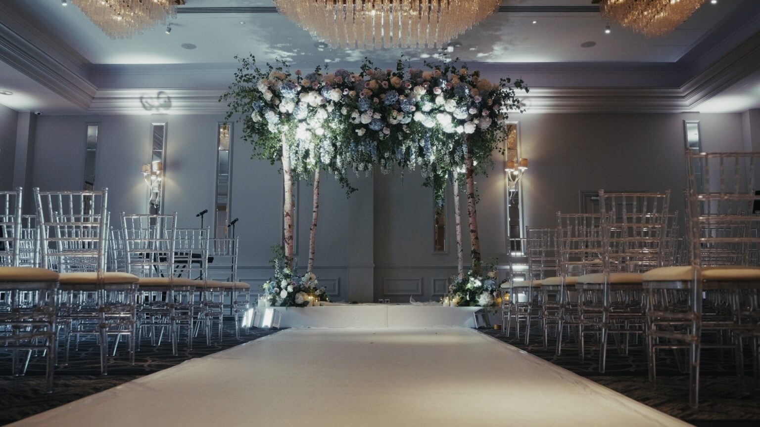A chuppah and white aisle runner beneath a floral canopy in a hotel ceremony room