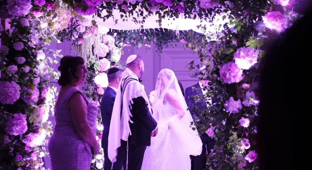 Wedding ceremony beneath a floral chuppah lit in violet, Just Smile Ltd