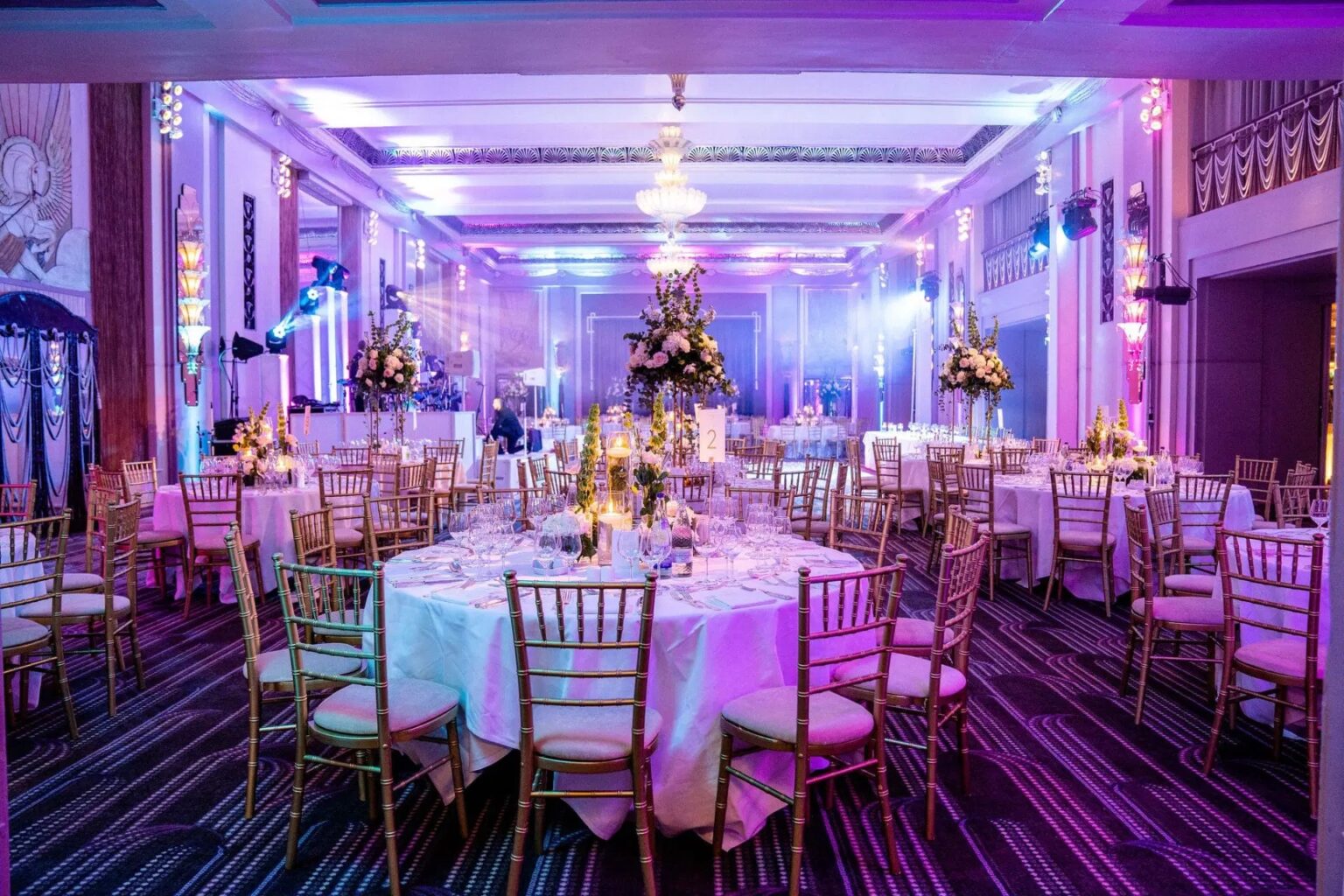 A banqueting room with round tables, uplighting and chandeliers