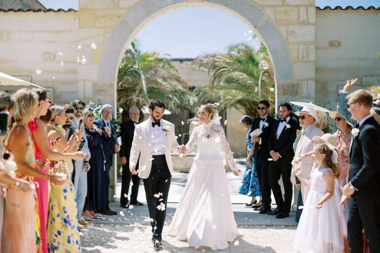 A bride in a long sleeved gown with guests at a stone archway, Kate Edmondson Bridal Couture