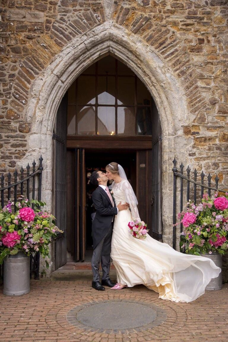A bride and groom kissing at a gothic church doorway, the train blown out behind her, Kate Edmondson Bridal Couture