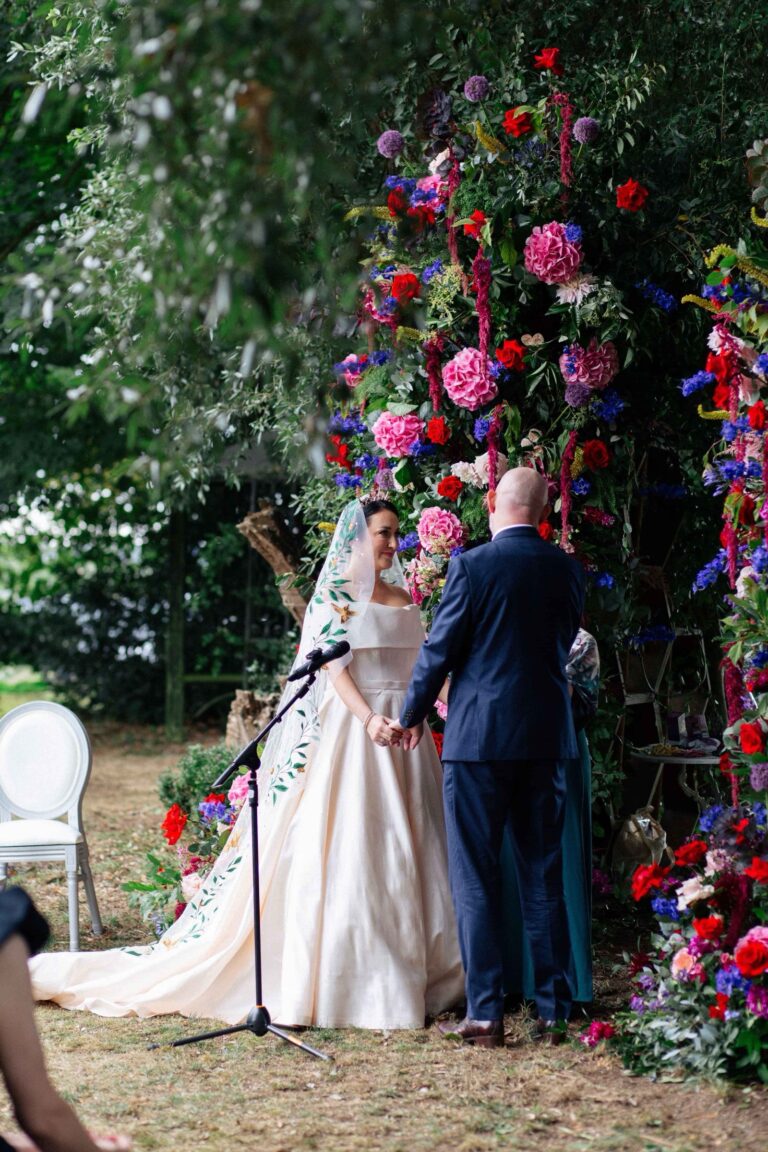 A bride in a long train gown at a ceremony beneath a flower arch, Kate Edmondson Bridal Couture