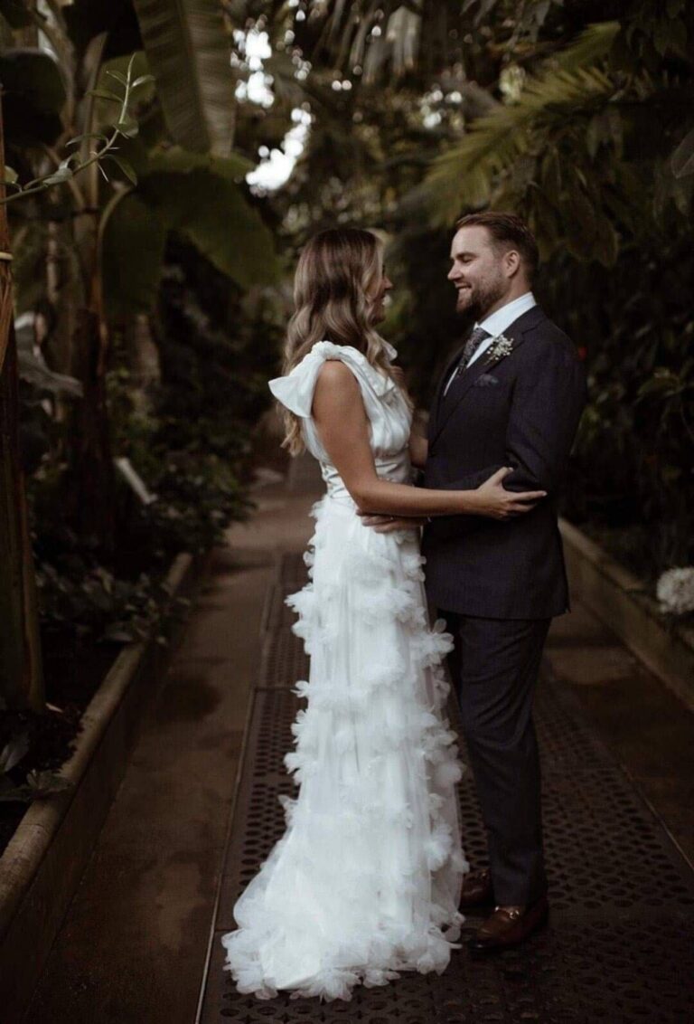 A bride and groom in a conservatory, her gown finished with tiered ruffles, Kate Edmondson Bridal Couture